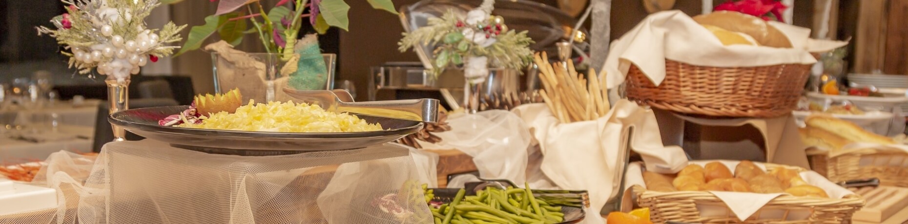 Buffet table with yellow food, green vegetables, bread rolls, and floral decorations, set for a festive gathering.