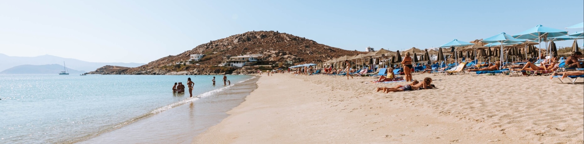Spiaggia serena con persone che si rilassano, nuotano e passeggiano, sotto un cielo blu e una collina rocciosa sullo sfondo.