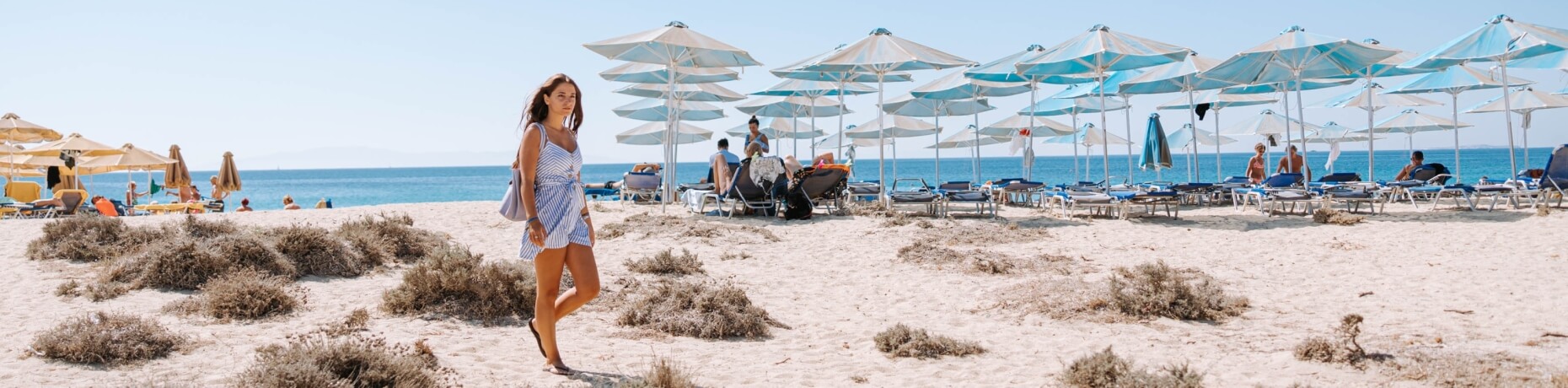 Una donna in abito a righe blu e bianche cammina su una spiaggia serena, circondata da ombrelloni e un mare tranquillo.