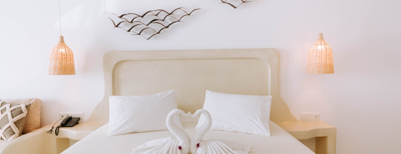 Bright hotel bedroom with a king bed, cream nightstands, curved sofa, and towel swans, filled with natural light.