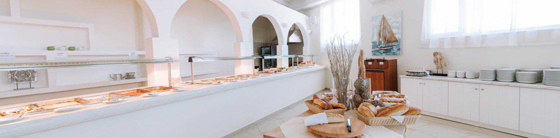 Un lungo tavolo buffet bianco con vari piatti, circondato da tavoli aggiuntivi in un ambiente luminoso e accogliente.