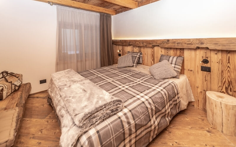 Camera accogliente con letto grande, coperta a quadri, panchina in legno e tende leggere, trasmette calore e comfort.