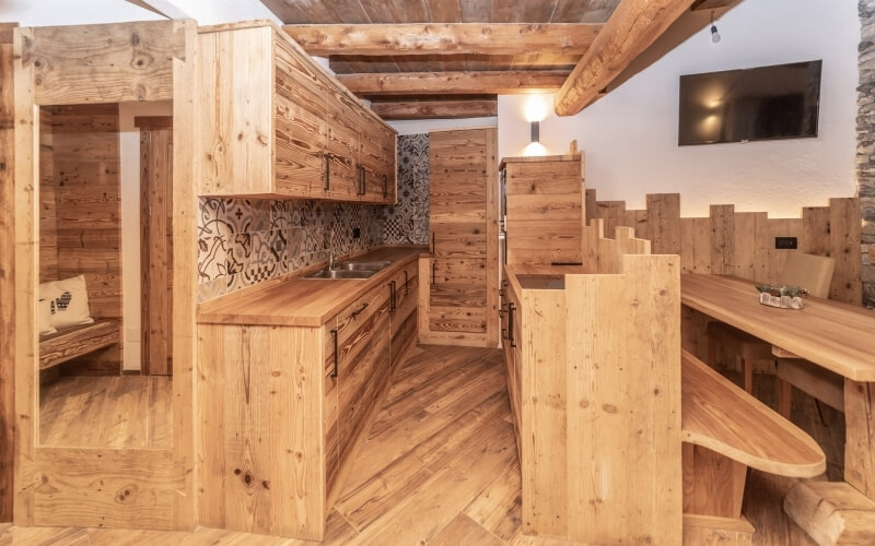 Cucina e sala da pranzo open space con isola centrale, panca, tavolo per sei e TV montata, in toni di legno chiaro.