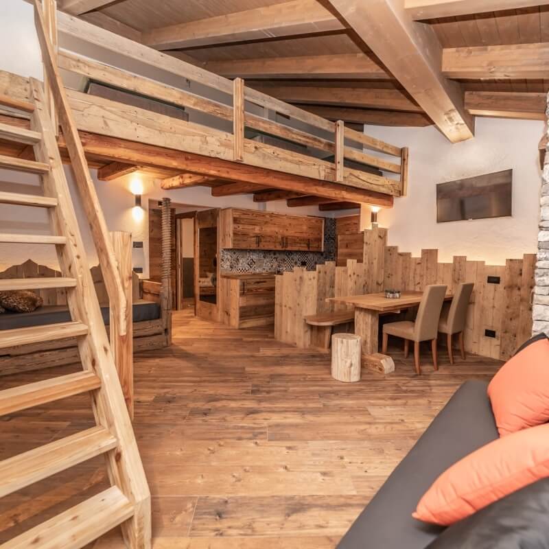 Rustic open-plan living space with wooden floors, loft area, modern sofa, dining table, and kitchenette. Cozy atmosphere.