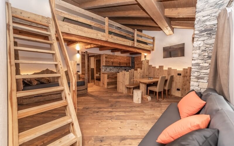 Rustic open-plan living space with a spiral staircase, loft bed, black couch, and dining area, featuring warm wood tones.