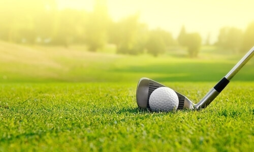 Close-up of a white dimpled golf ball on grass with a sleek golf club partially visible, set against a blurred serene landscape.