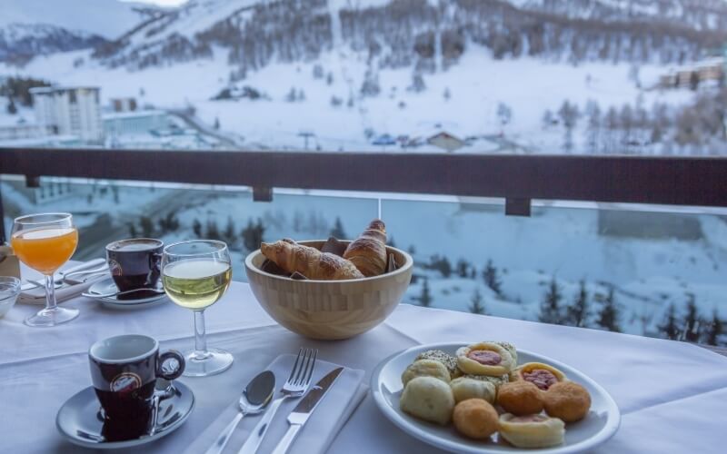 Tavolo da colazione su un balcone con vista su un paesaggio innevato, con cibo e bevande vari.