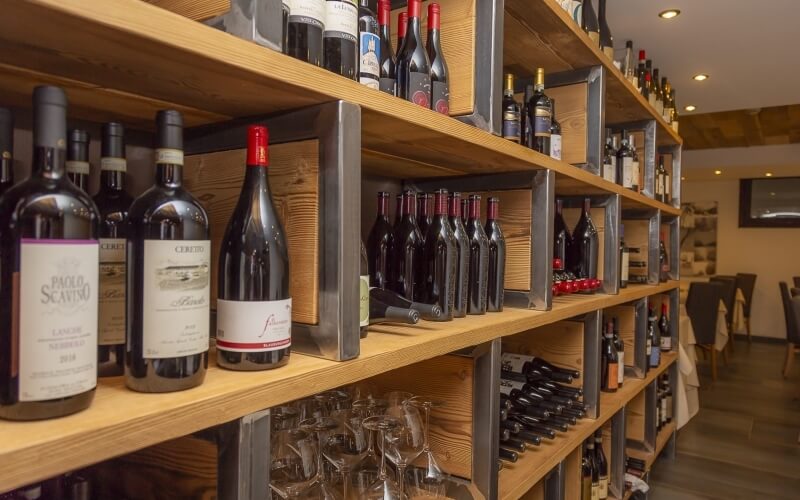 Wine bottles on wooden shelves, with glasses below and a blurred room with a table and TV in the background.