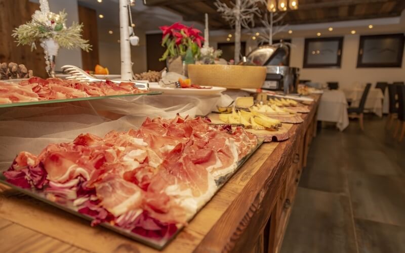 Buffet festivo con prosciutto, formaggio, decorazioni natalizie e un'area pranzo elegante sullo sfondo.