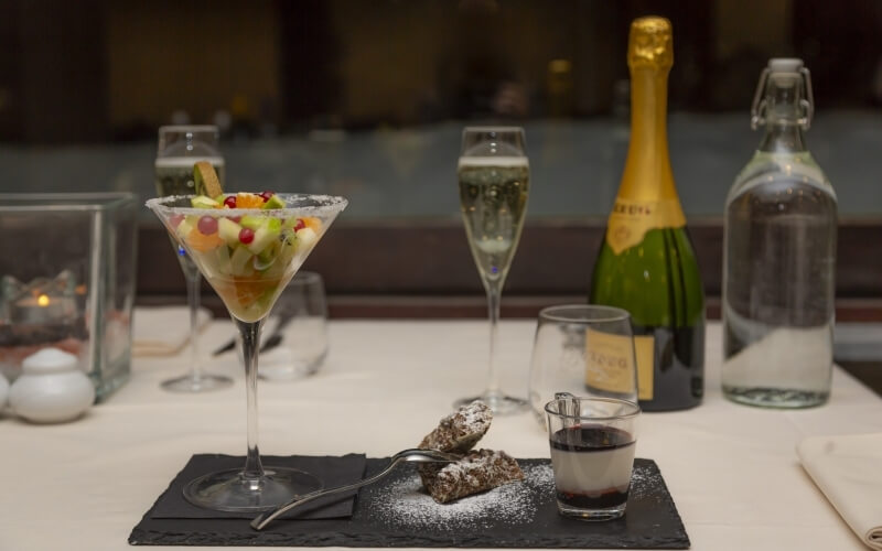 Tavolo elegante per dessert con martini di frutta, biscotti al cioccolato, vino bianco e champagne, illuminato da una candela.