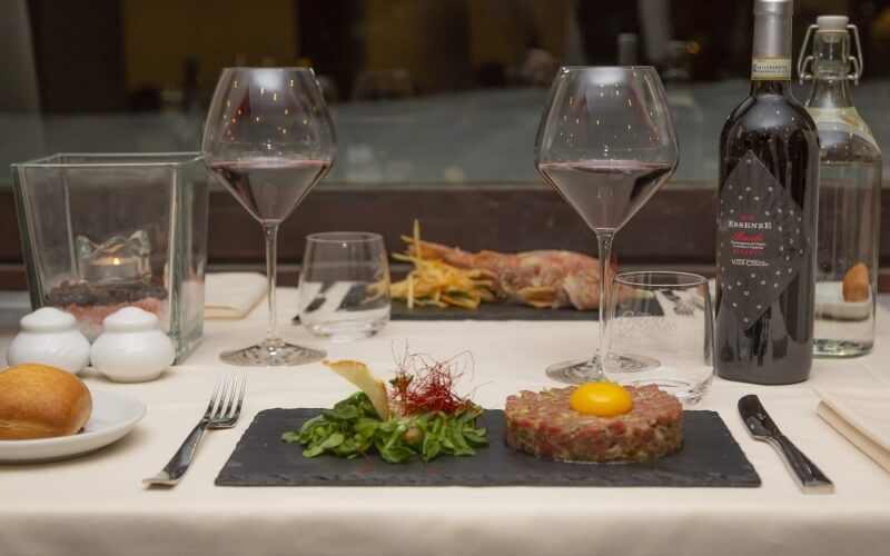 Elegant dining table set with steak tartare, wine, and condiments, featuring a white tablecloth and sophisticated decor.