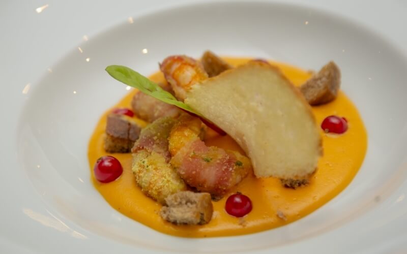 Close-up of a white bowl with yellow sauce topped with prawns, croutons, breadcrumbs, cranberries, and a green leaf.