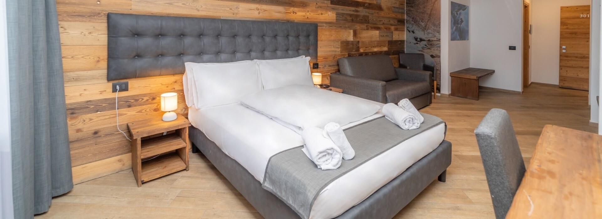 Cozy hotel room with king-size bed, nightstand, couch, and light wood paneling, featuring a doorway to a bathroom and hallway.