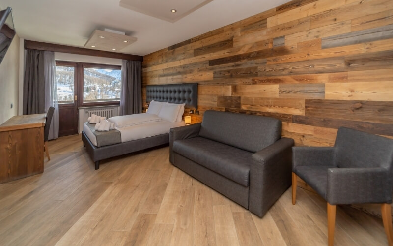 Cozy hotel room with rustic wood-paneled wall, bed, couch, chairs, and desk, creating a warm and inviting atmosphere.