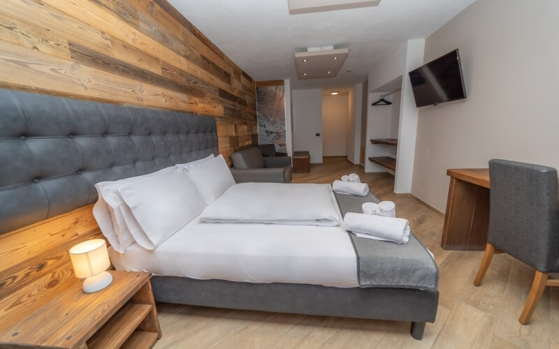 Cozy hotel room with a bed, nightstand, desk, and gray chair; features wooden accents and an open doorway to another room.