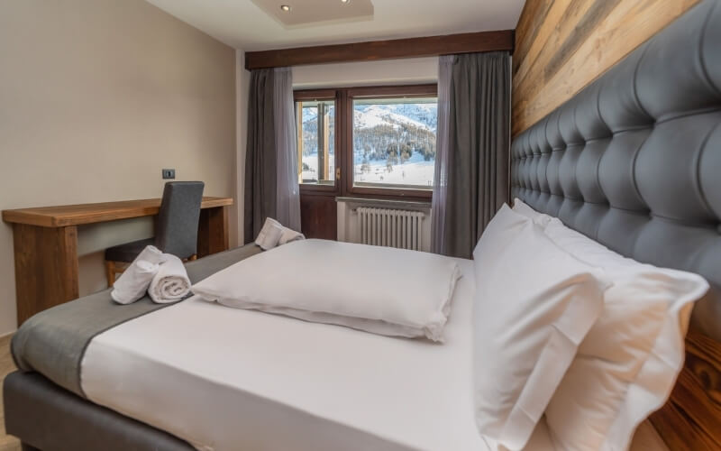 A cozy bedroom with a gray tufted bed, wooden desk, and a window showcasing snowy mountains outside.