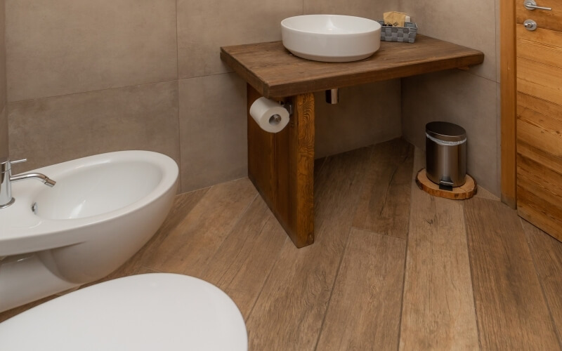 A well-maintained bathroom featuring a white bathtub, wooden table with sink, toilet, tan tiled walls, and wooden floor.