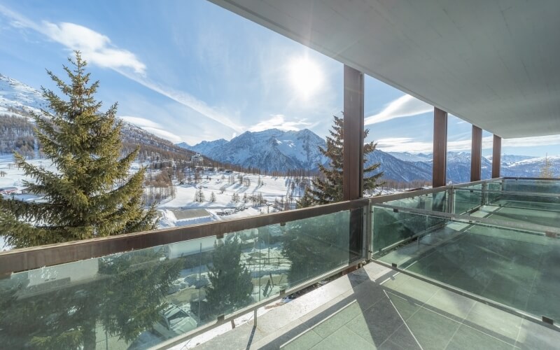 Balcony with glass railing overlooking snow-covered mountains and trees under a blue sky with clouds.