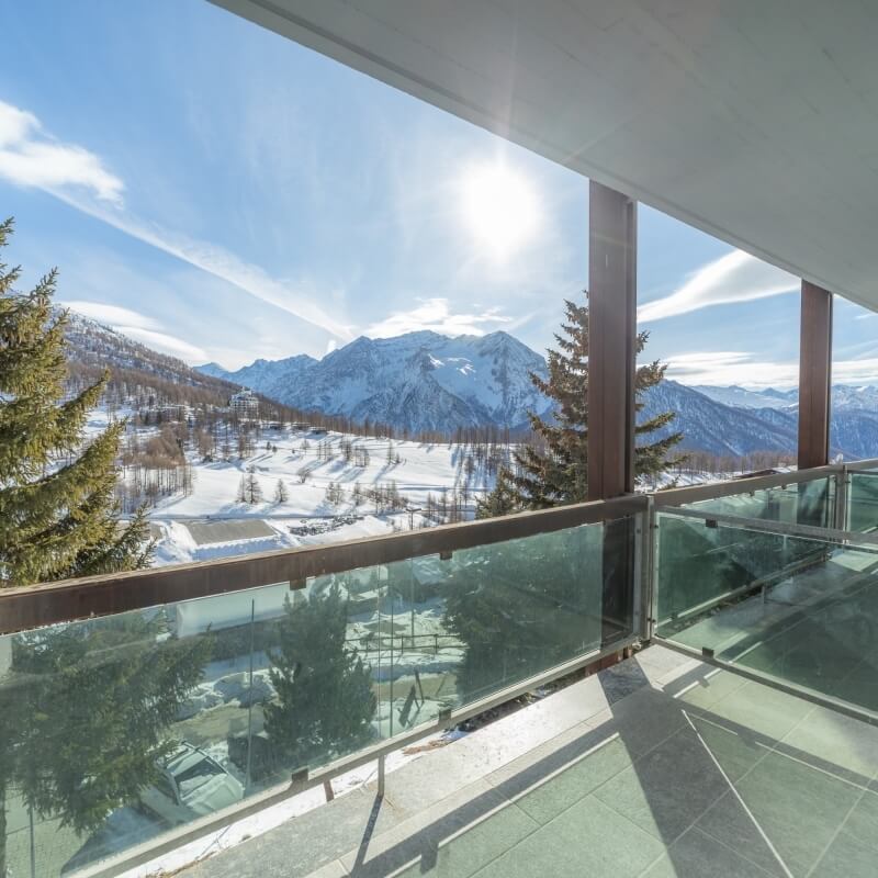 Serene winter scene from a balcony, featuring snow-covered mountains, evergreen trees, and a bright blue sky.