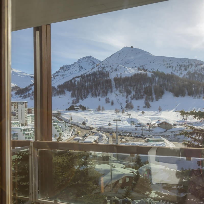 Serene winter scene through a large window, featuring snow-covered mountains, a ski lift, and reflections of trees and buildings.