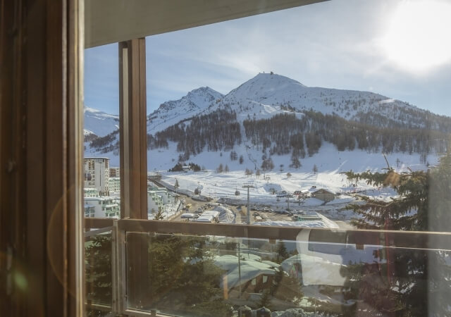 Serene winter landscape featuring snow-covered mountains, trees, and buildings, viewed from a building interior with sunlight.