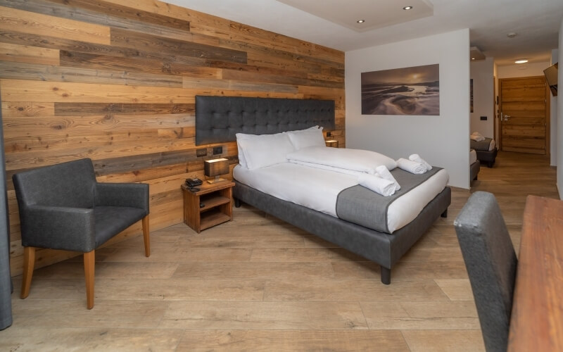 Modern hotel room featuring a gray bed, chair, nightstand with lamp, and beach photograph on the wall, with light wood flooring.