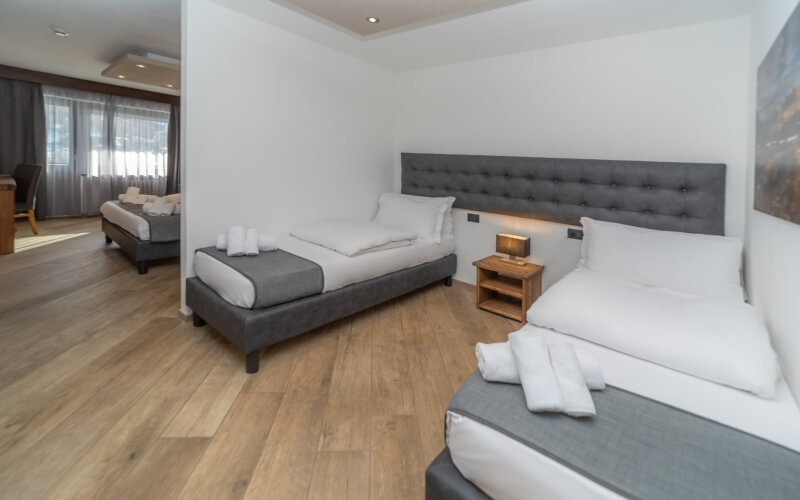 Modern hotel room with two beds, nightstand, and adjacent room visible; minimalist design with light wood floors and white walls.