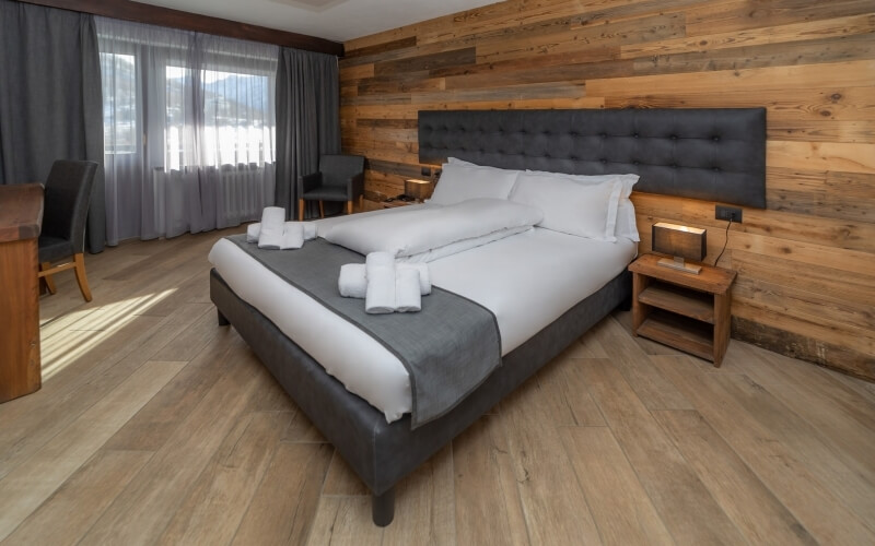 Cozy hotel room with a gray bed, wooden decor, two chairs by a window, and a small desk, featuring natural light.