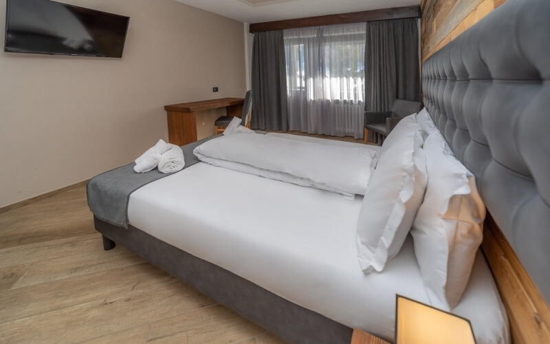 Hotel room featuring a large bed, desk, window with curtains, flat-screen TV, and light wood flooring in neutral tones.