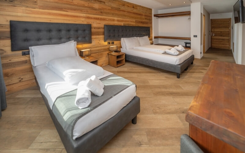 Modern hotel room with two beds, a nightstand, wooden desk, and mounted TV, featuring light wood floors and white paneling.