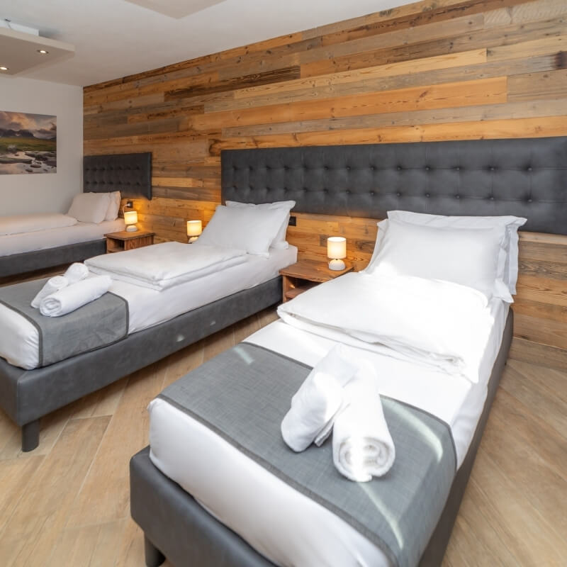 Cozy hotel room with three beds, dark gray bases, white linens, wood-paneled walls, and warm lighting.