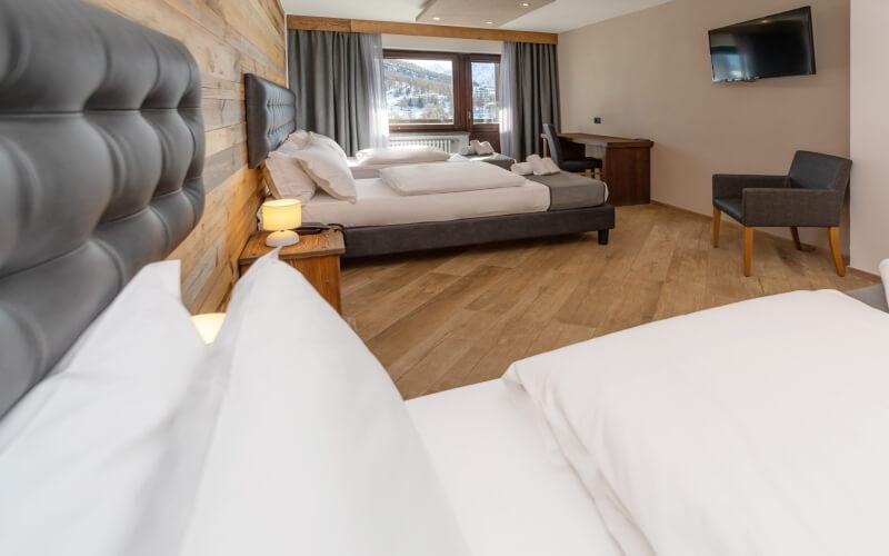 Camera d'hotel con due letti, pavimento in legno chiaro, finestra con tende grigie e TV montata sulla parete.