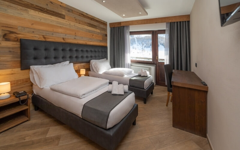 Hotel room with two beds, wooden accent wall, large window with snow view, desk, nightstand, and neutral decor.