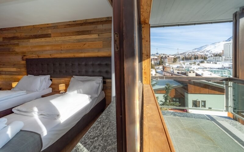 Modern hotel room with two beds, balcony view of the city, wood paneling, and a serene blue sky.