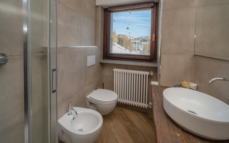 Modern bathroom with beige tiles, wooden floor, glass shower, sink, and cityscape view through a window.