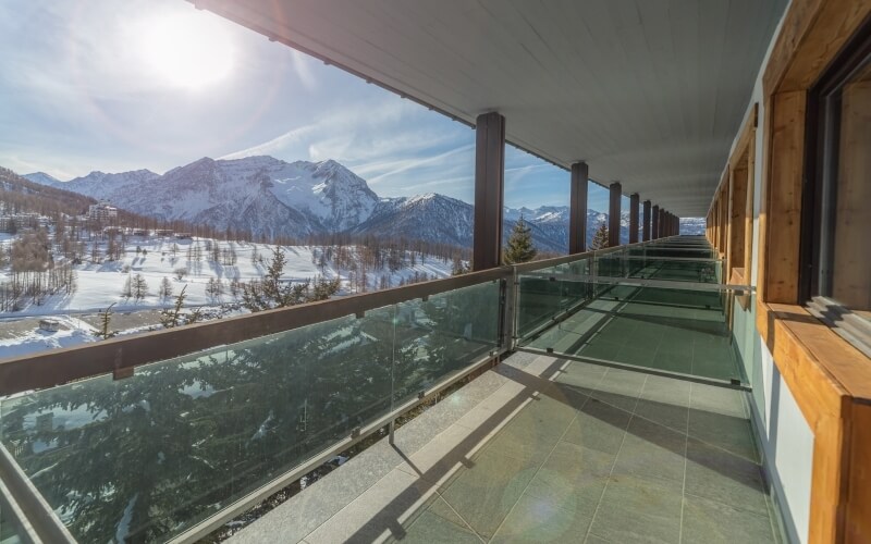 Balcony with glass railing overlooking snow-covered mountains and a serene valley under a blue sky with wispy clouds.