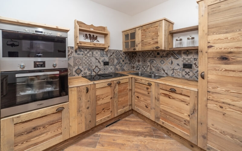 Angolo di cucina con armadi in legno, forno in acciaio inox, piano nero e pareti in piastrelle grigie e bianche.