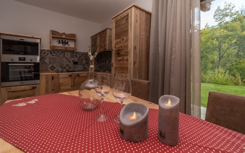 Tavolo da pranzo per due con runner a pois, candele, calici, vista su un prato verde e cucina sullo sfondo.