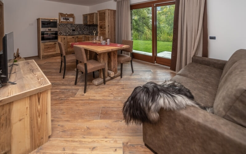 Cozy living room and dining area with a brown couch, wooden table, and large window overlooking a lush patio and lawn.