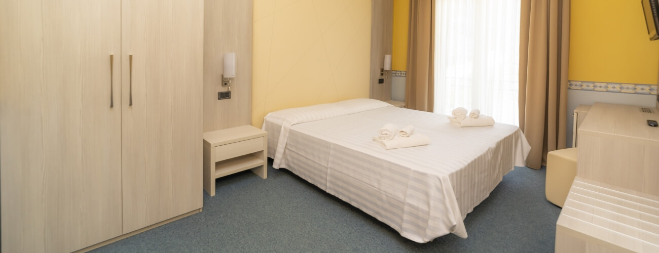 Camera d'hotel con letto centrale, armadio, tavoli, finestre con tende e pareti gialle e beige. Atmosfera pulita e confortevole.