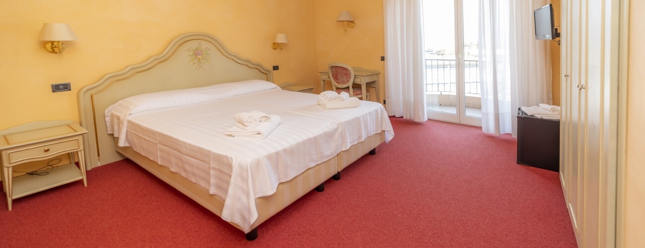 Camera d'albergo con letto, comodini, sedia, TV, tappeto rosso e pareti gialle, con porta scorrevole verso un balcone.