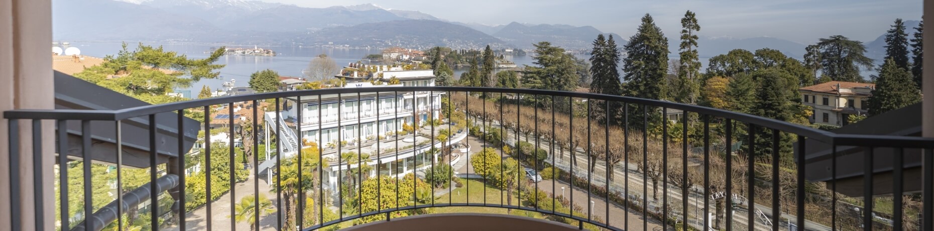 Balcony view of a serene lake, mountains, and a charming town, framed by lush greenery and a white multi-story building.