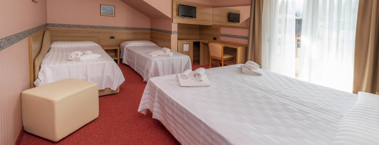 Cozy hotel room with four beds, pink walls, red carpet, desk, and window with sheer curtains letting in natural light.