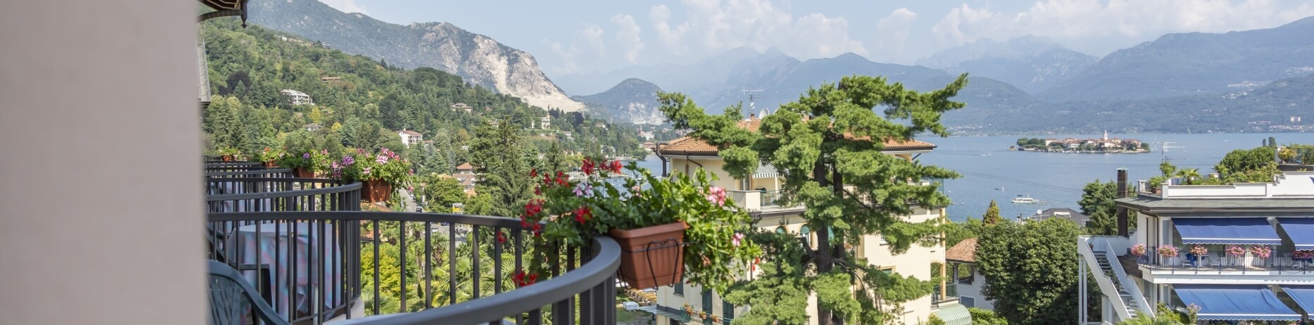 Serene balcony with potted plants, overlooking a picturesque town and mountains under a blue sky with fluffy clouds.