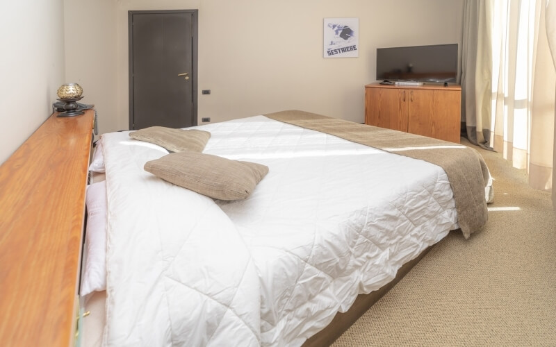 A well-lit minimalist bedroom featuring a large bed, TV on a dresser, and sheer-curtained windows.