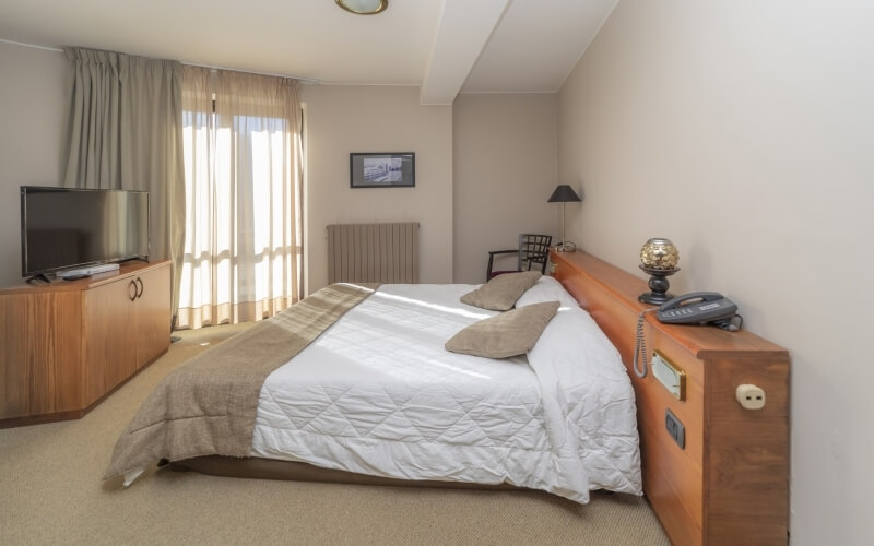 A cozy hotel room featuring a bed, nightstand, television, dresser, and window with sheer curtains, all in warm tones.