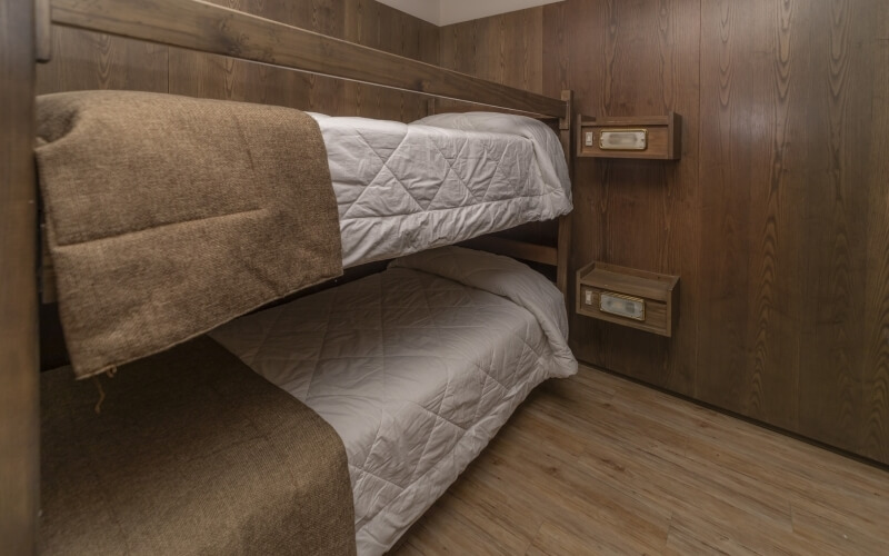 Rustic small room with L-shaped bunk beds, white bedding, brown blankets, wooden shelves, and light hardwood floor.