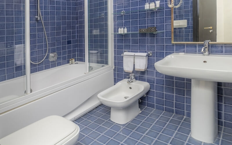 A serene blue-tiled bathroom featuring a bathtub, bidet, pedestal sink with a gold-framed mirror, and minimalist decor.