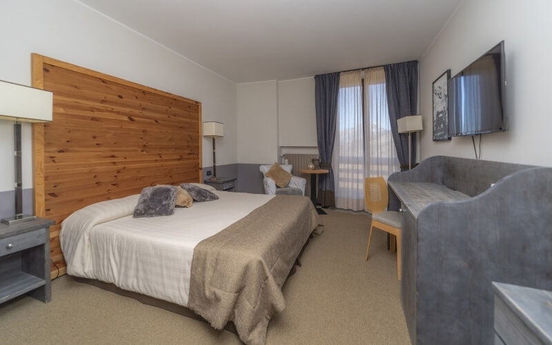 Camera d'hotel con letto, comò, TV, decorata in toni neutri, con luce naturale che entra da una grande finestra.