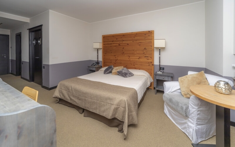 Hotel room with a central bed, gray nightstands, white couch, and tan carpet, exuding simplicity and comfort.